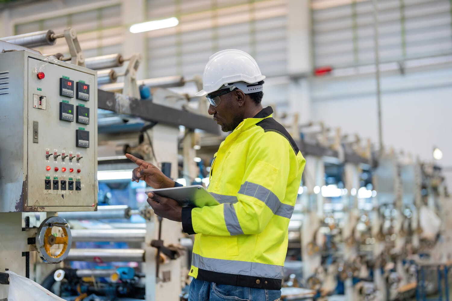 An operator in a high-visibility jacket checks a machine with a tablet on the production floor